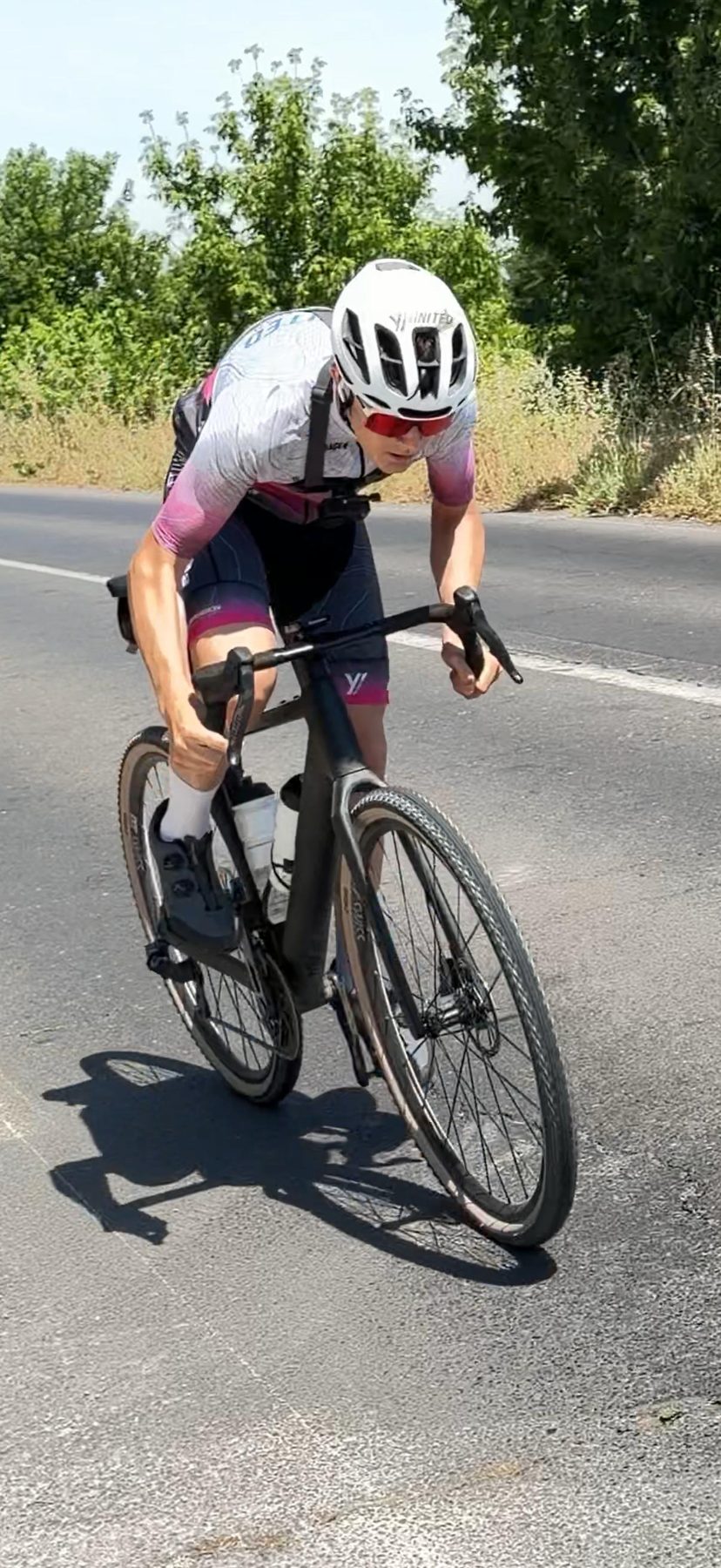 Fahrer im Teamtrikot auf dem Rennrad auf einer schmalen Landstraße zwischen Feldern.