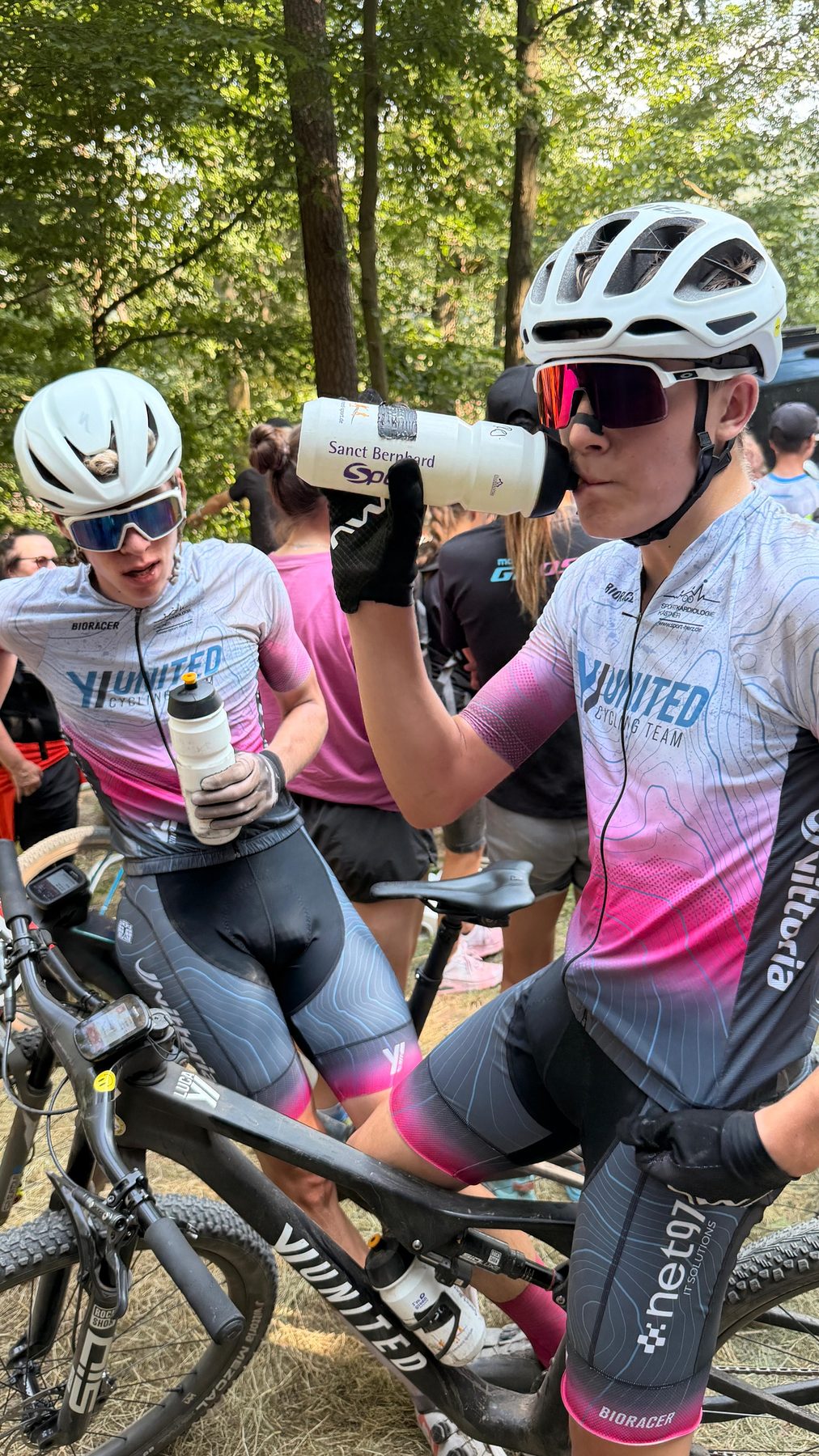 Zwei Fahrer im Teamtrikot trinken nach dem Rennen aus ihren Trinkflaschen, hinter ihnen der Wald und weitere Aktive.