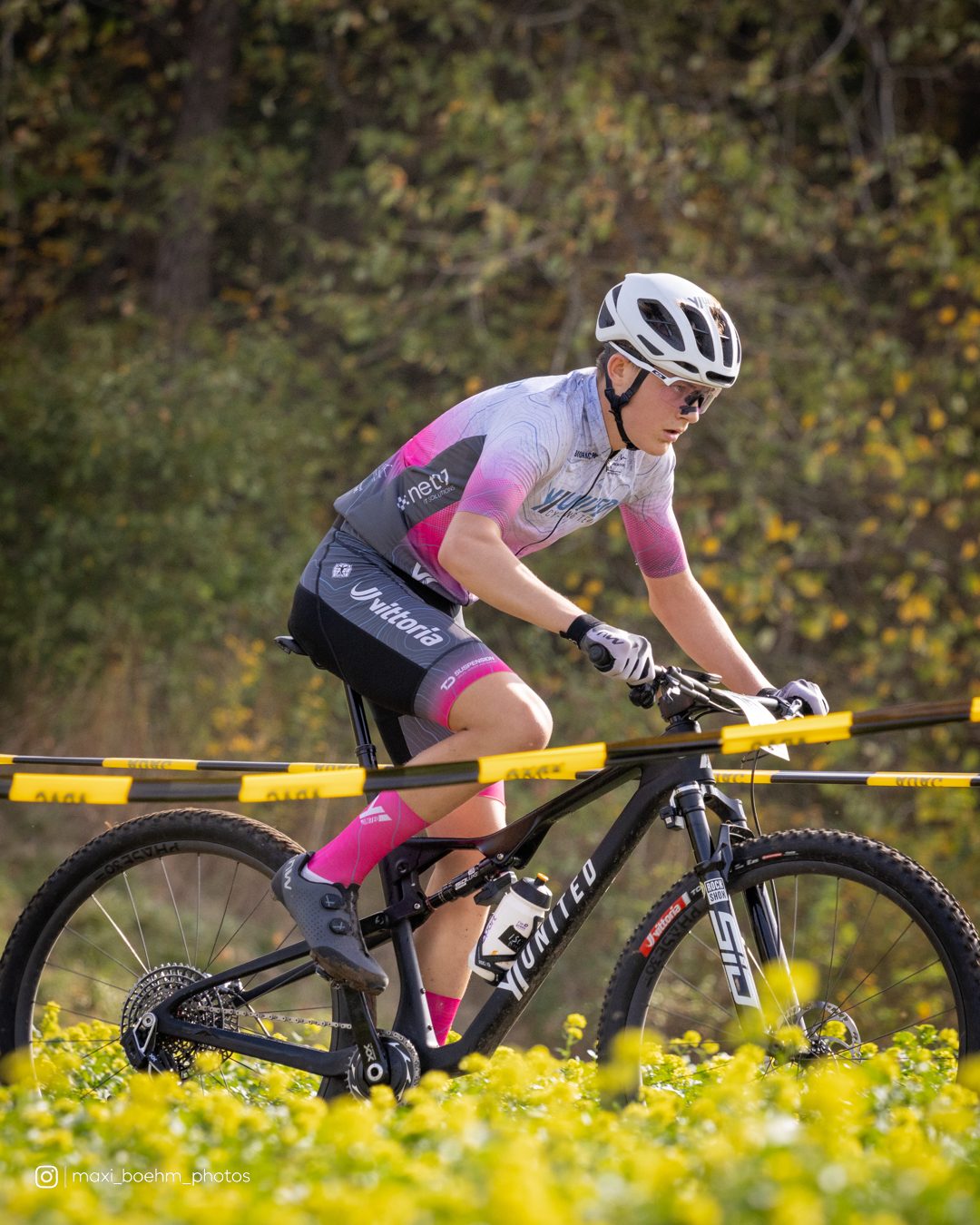 Fahrer im weiß-rosa Teamtrikot fährt an einem blühenden Rapsfeld entlang, hinter ihm die gelbe Streckenabsperrung.