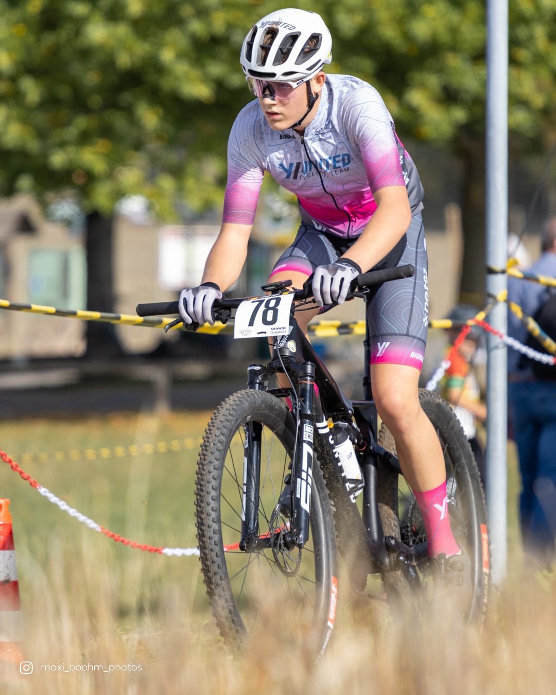 Fahrer im weiß-rosa Teamtrikot mit der Startnummer 78 tritt an einer abgesperrten Wiese entlang, im Hintergrund Bäume und Zuschauer.