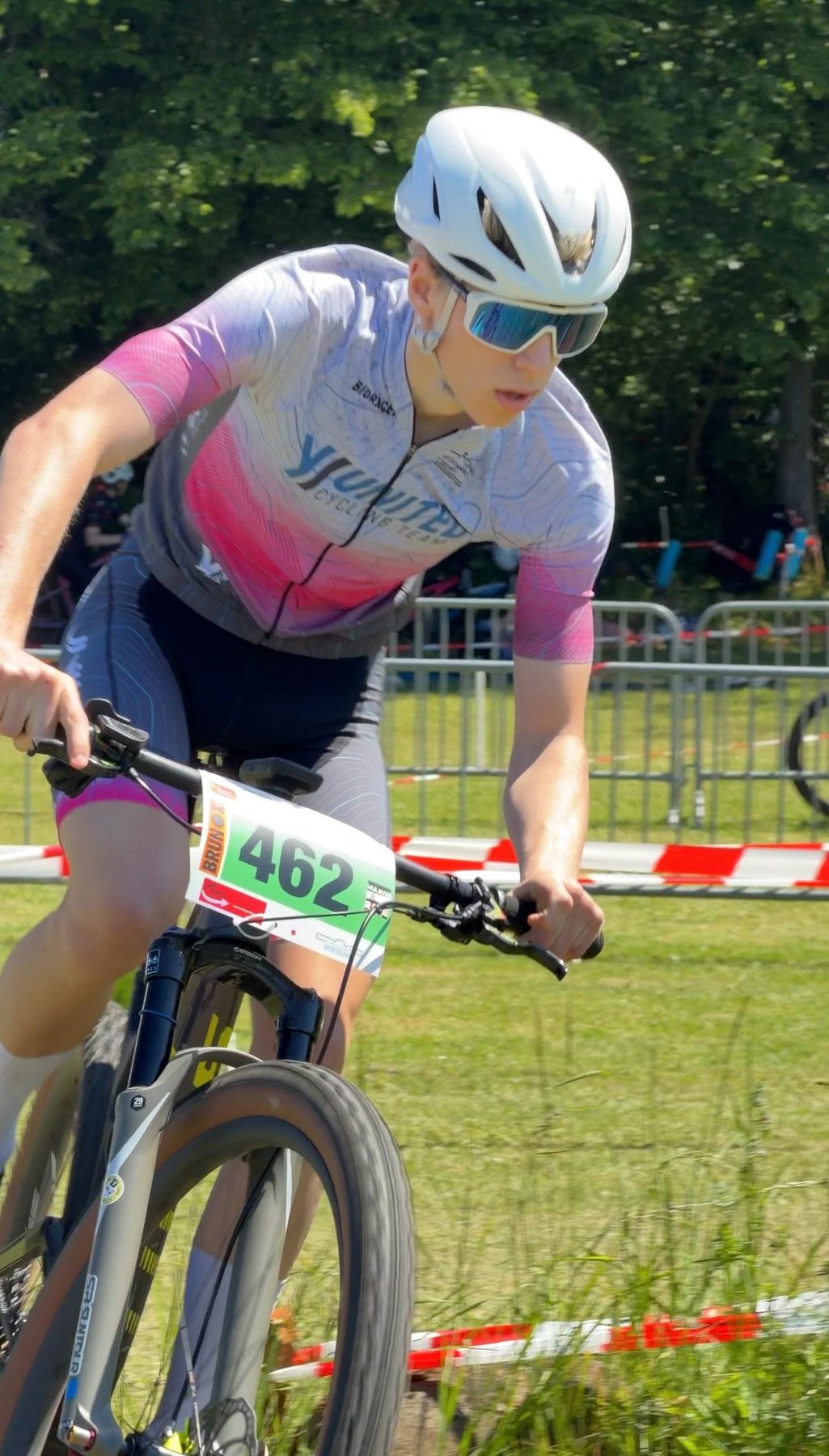 Fahrer mit der Startnummer 482 im weiß-rosa Teamtrikot tritt im Rennen über eine Wiese, am Rand flattert Absperrband.