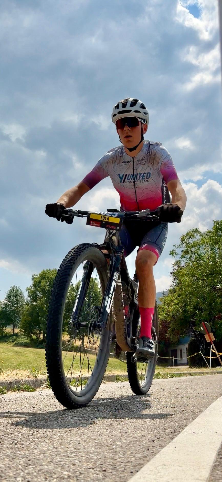 Fahrer im Teamtrikot rollt über einen asphaltierten Weg, im Hintergrund Bäume und Wiese.